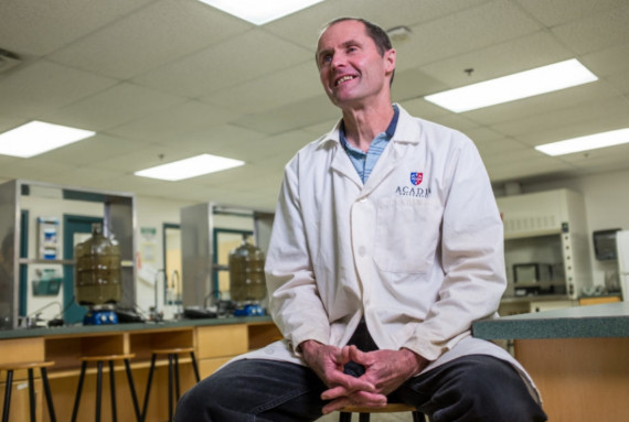 Man in a white lab coat sitting on a chair smiling.